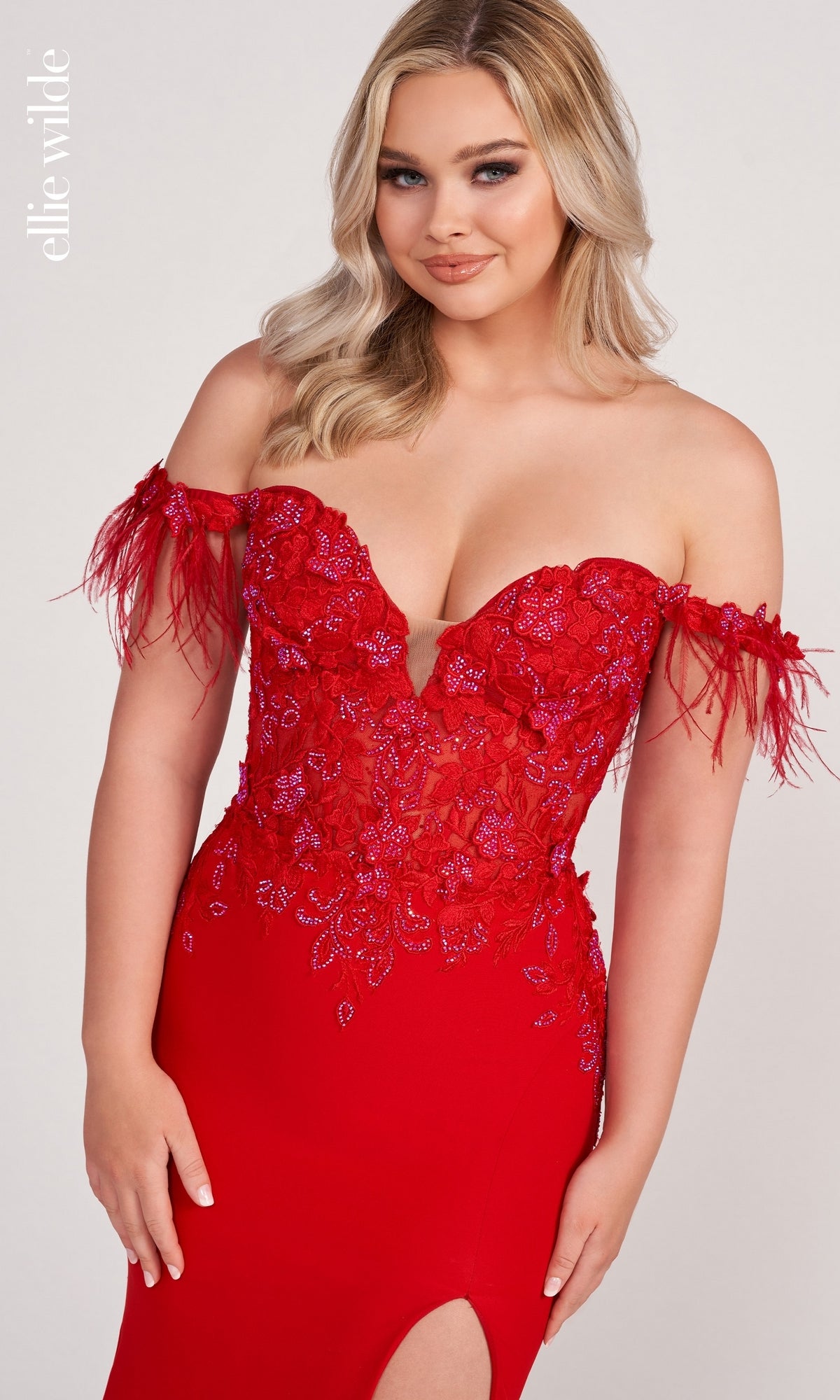A woman stands against a light background wearing the Ellie Wilde Feathered Off-Shoulder Long Prom Dress, featuring red floral lace and feathered sleeve details.